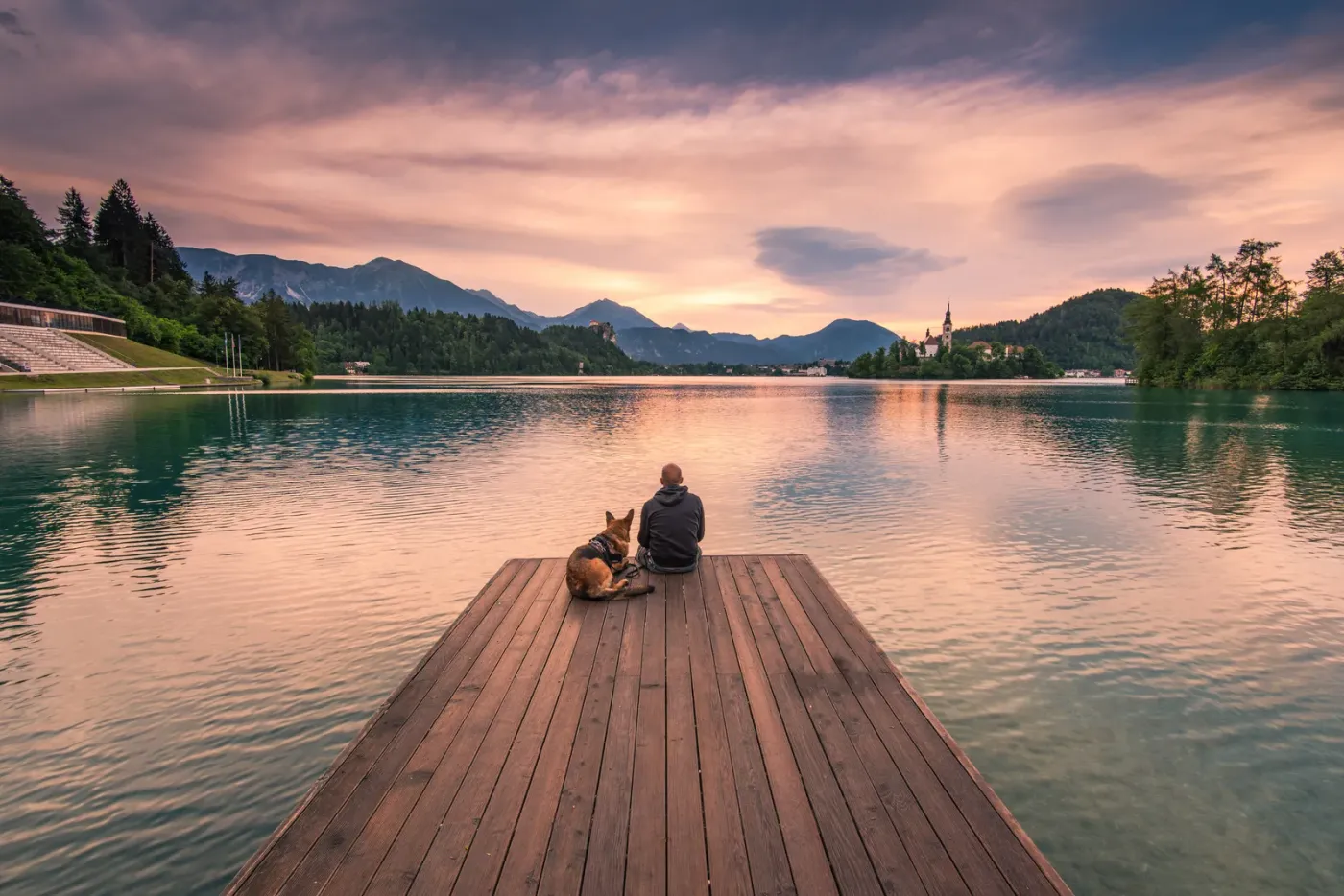 Person and dog sitting on a dock at a mountain lake at sunset