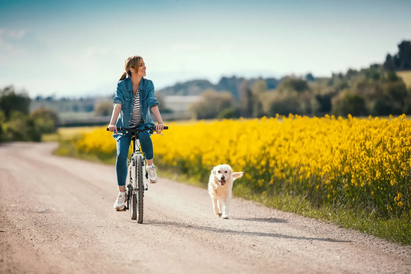 Woman cycling a gravel road beside a canola field with her dog running alongside