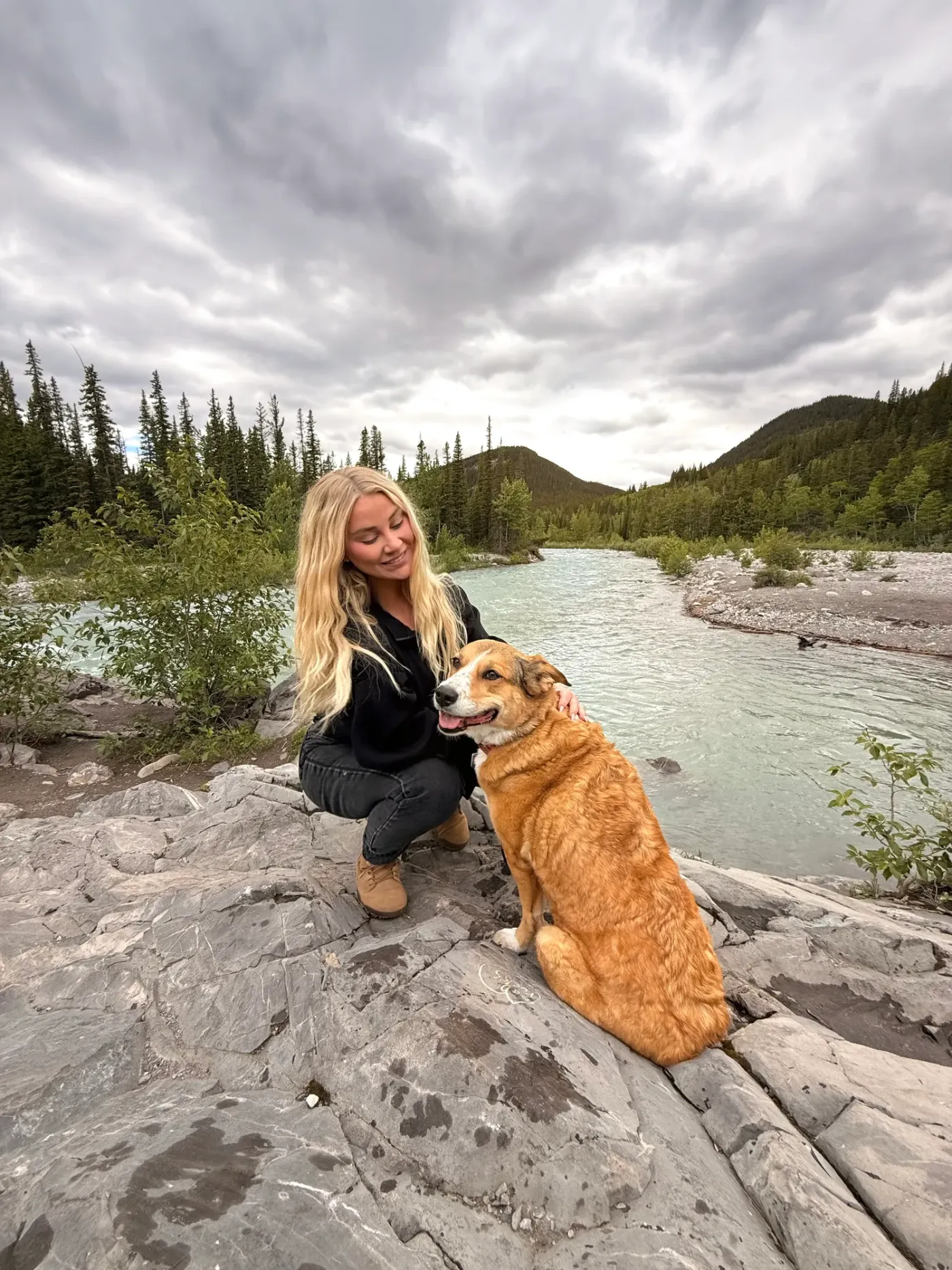 Trainer and a golden cross sitting together on riverside rocks