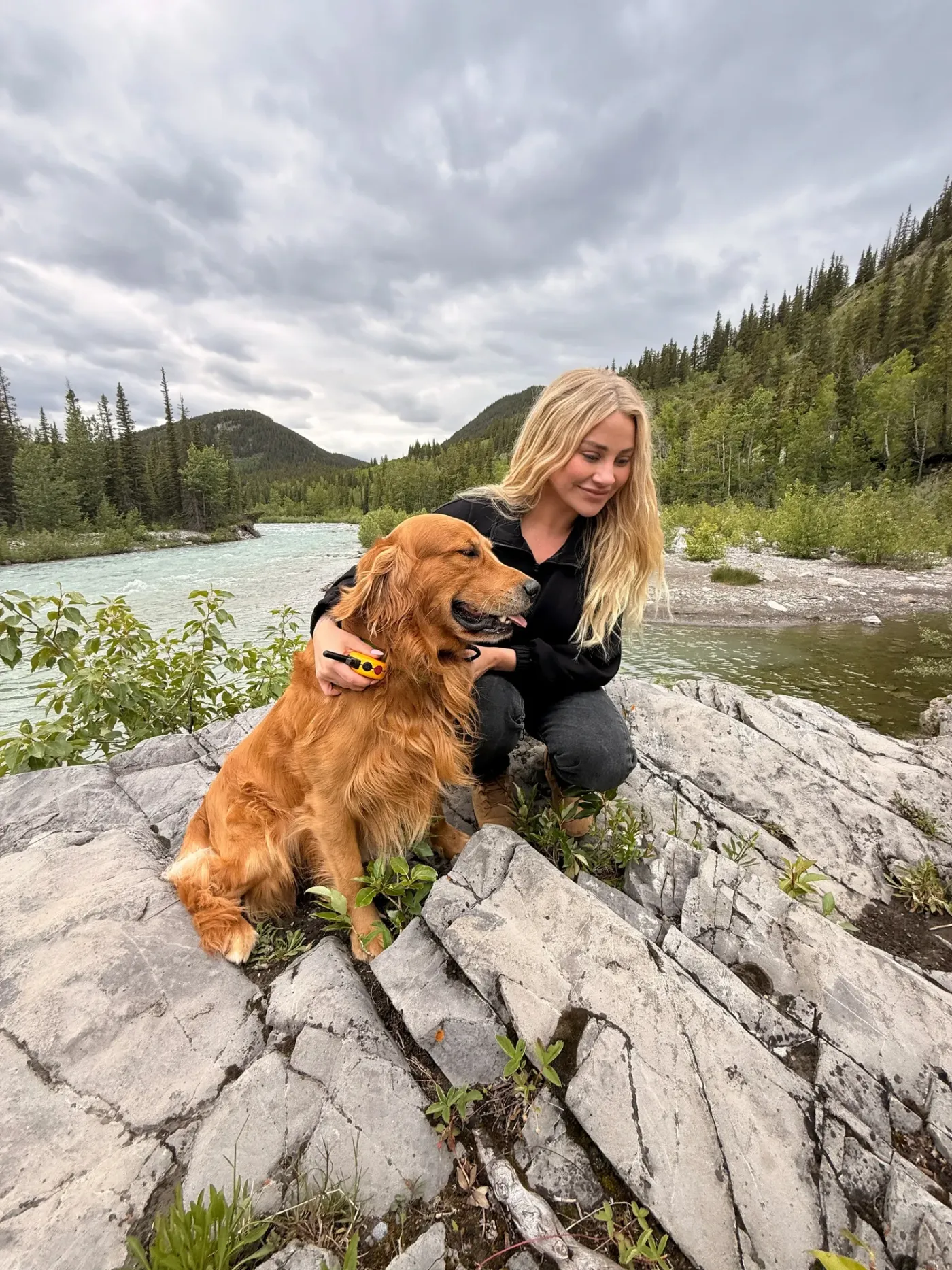 Trainer crouched beside a golden retriever on the rocks of a mountain river